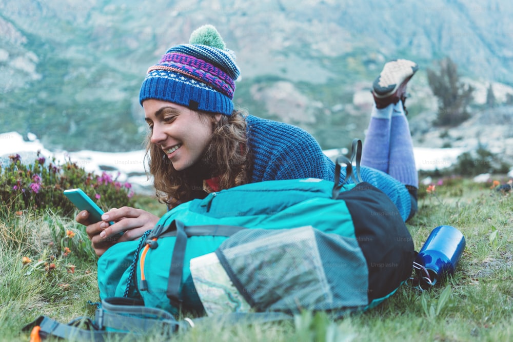 Young happy traveling woman with backpack, hat lying in the grass and flowers and using mobile phone, smilling in the stunning mountain wilderness after day of hiking.