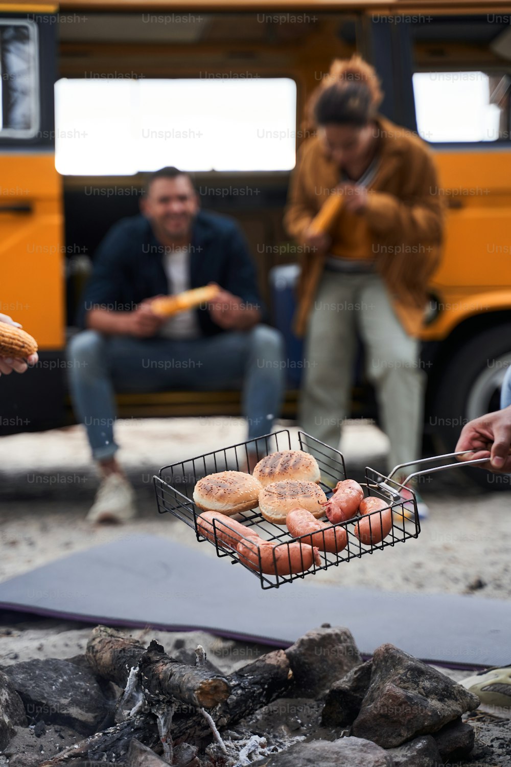 Group of tourists preparing their lunch in the forest. Focus on sausage and bun on the grill. People are blurred