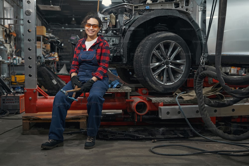 Trabajadora con gafas de seguridad sosteniendo martillos y sonriendo mientras descansa cerca del automóvil en el garaje
