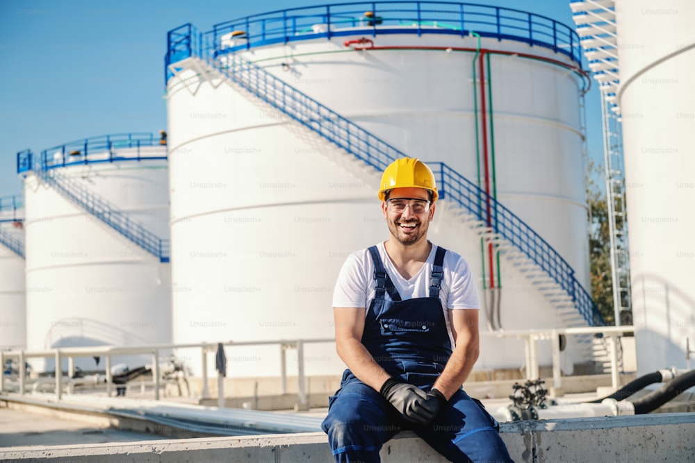 Cheerful caucasian worker in overall and with helmet on head sitting outdoors. In background are oil reservoirs.