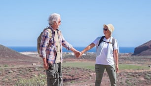 Happy active senior couple hiking in mountain enjoying healthy lifestyle and sunny day. Two elderly people hand in hand smiling, horizon over water. Vacation and sport concept