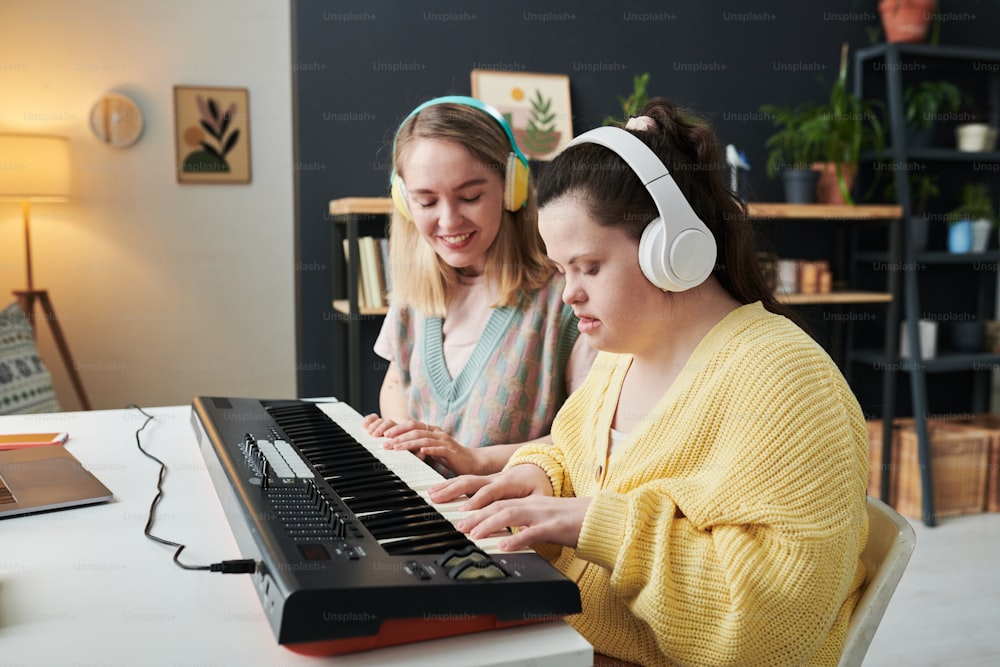 Joven mujer caucásica con auriculares que enseña a una niña con síndrome de Down a tocar el sintetizador en casa