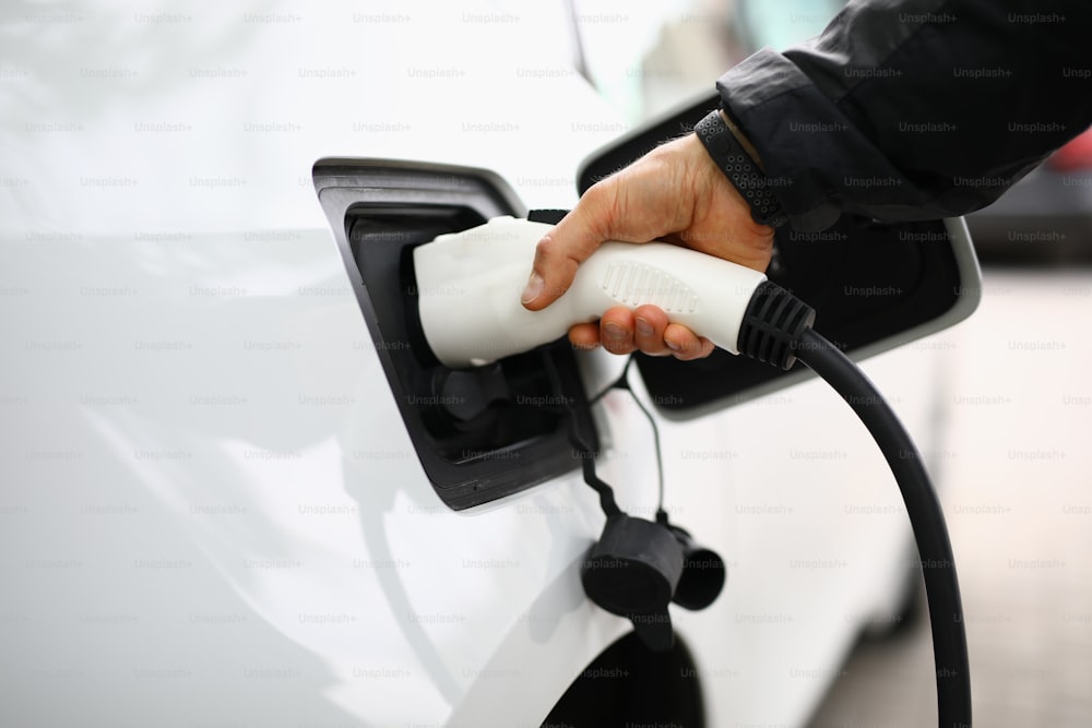 Male hand insert electric white gun into car. White electric car be recharged at charging station.