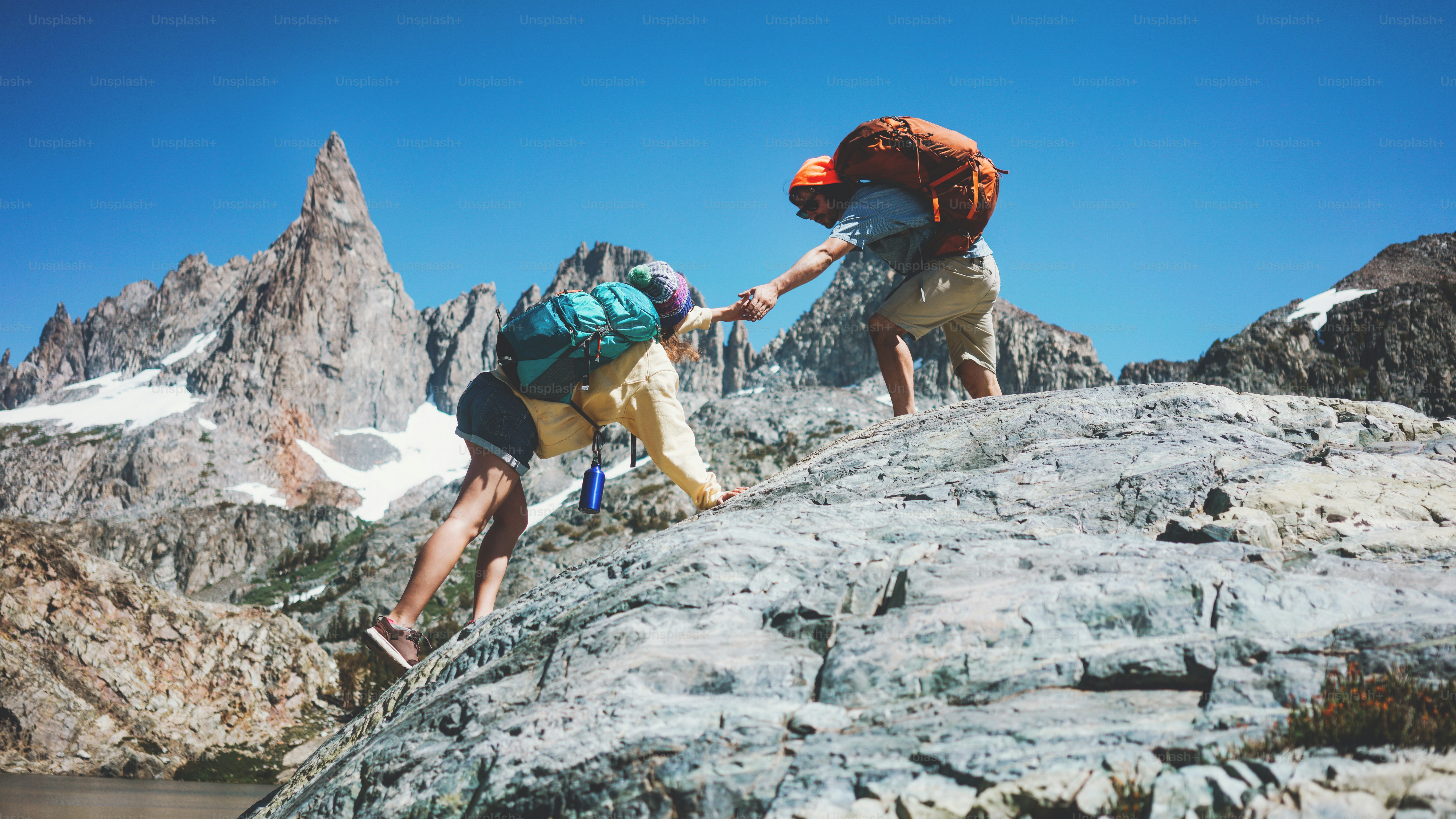 Young active couple with backpacks climbing rocks together in stunning snowy mountains. Natural wild near the lake