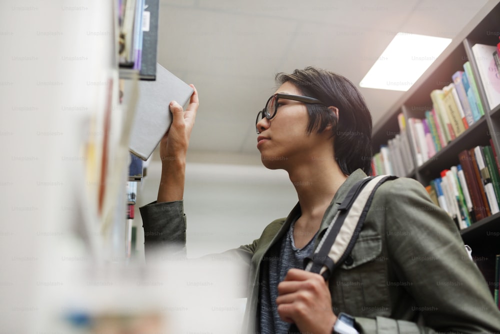 Asian young student in eyeglasses standing near the bookshelf and taking book he is in the library