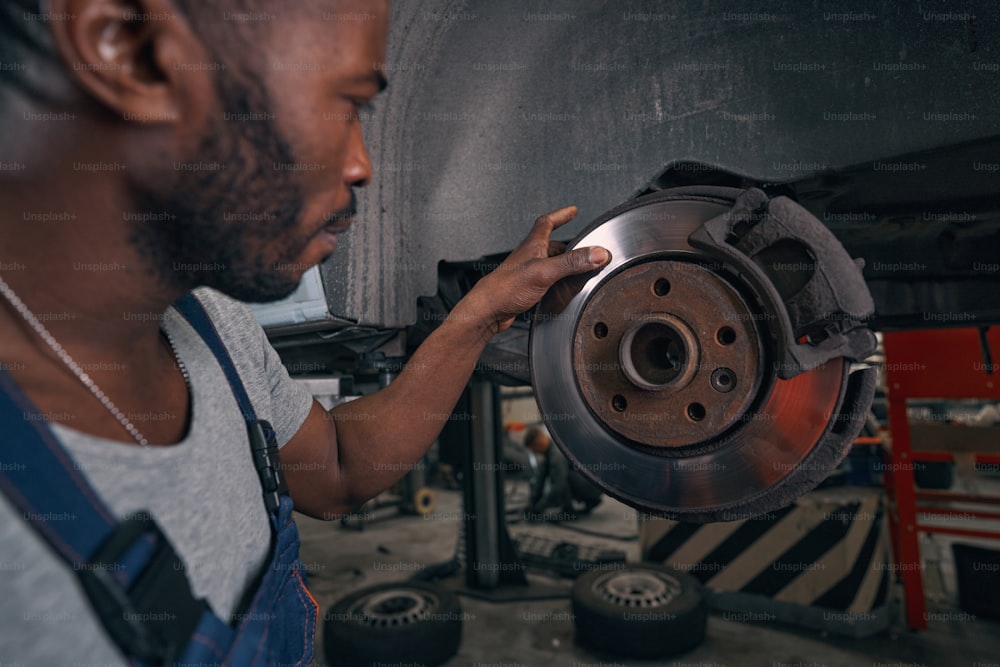 Männlicher afroamerikanischer Mechaniker, der während der Inspektion mit dem Finger über die Bremsscheibe des Autorades streicht