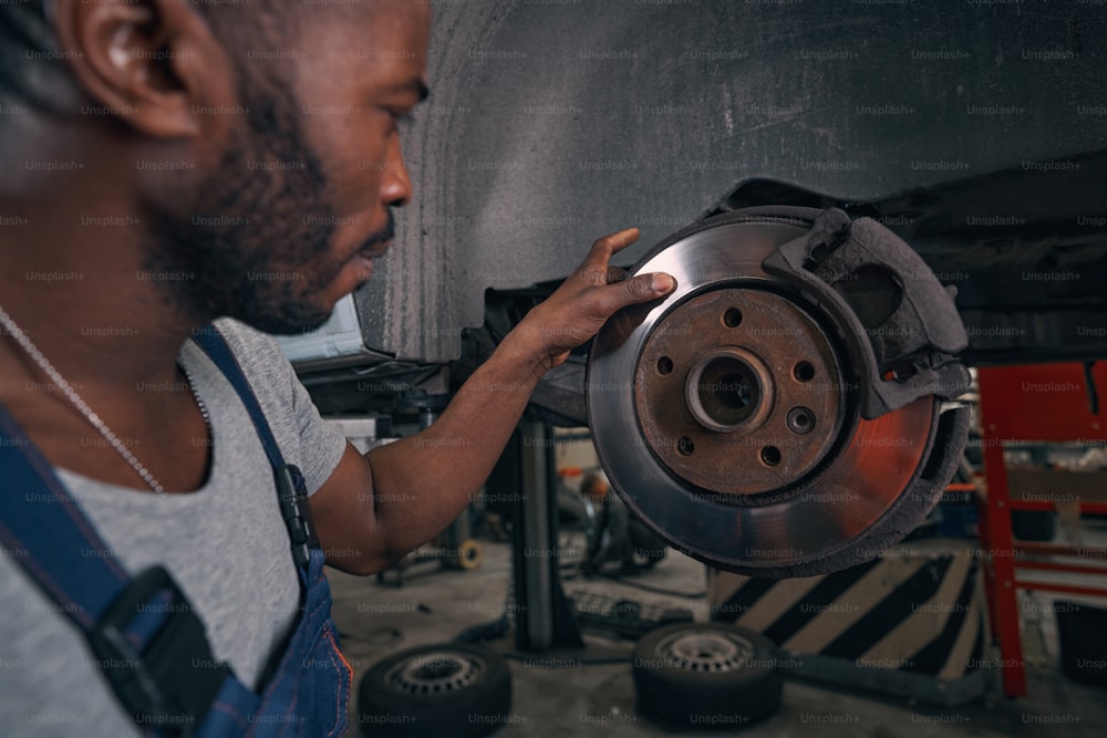 Male Afro-American mechanic sweeping his finger over brake disk of car wheel during inspection