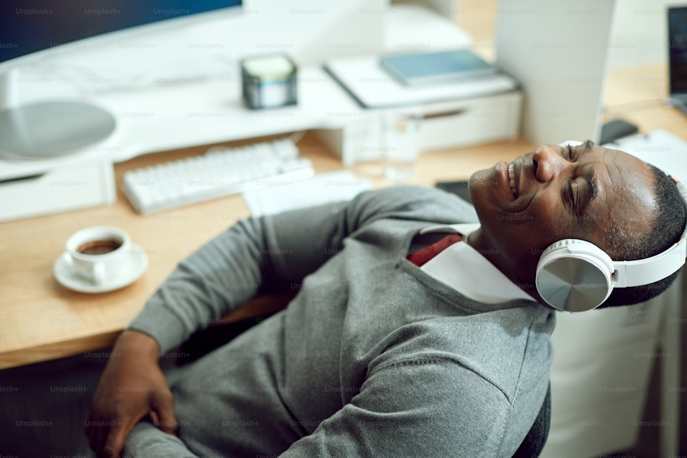 Smiling African American businessman listening music over headphones while relaxing with eyes closed in the office.