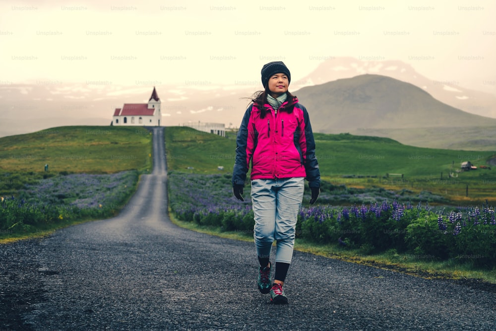 Woman traveler travel to Ingjaldsholl church in Hellissandur, Iceland in the field of blooming lupine flowers with background of Snaefellsjokull mountain. Beautiful sunny scenery of summer in Iceland.
