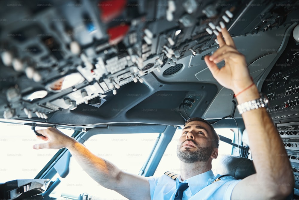 Pilota caucasico concentrato in uniforme seduto in cabina di pilotaggio e toccando gli interruttori sopra la sua testa