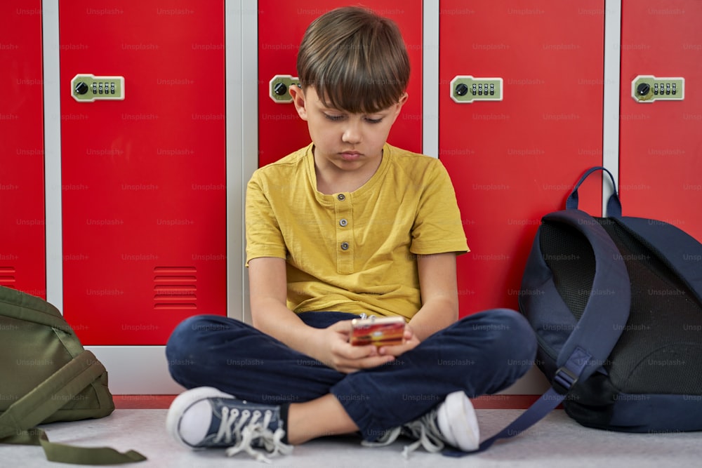 Sad schoolboy using phone and sitting against locker in school