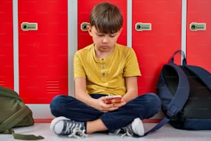 Sad schoolboy using phone and sitting against locker in school