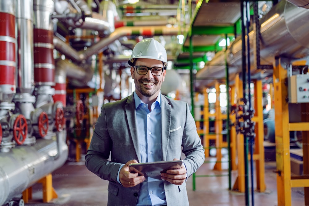 Beau superviseur souriant en costume gris et avec un casque blanc sur la tête tenant une tablette tout en regardant la caméra. Intérieur de la centrale.