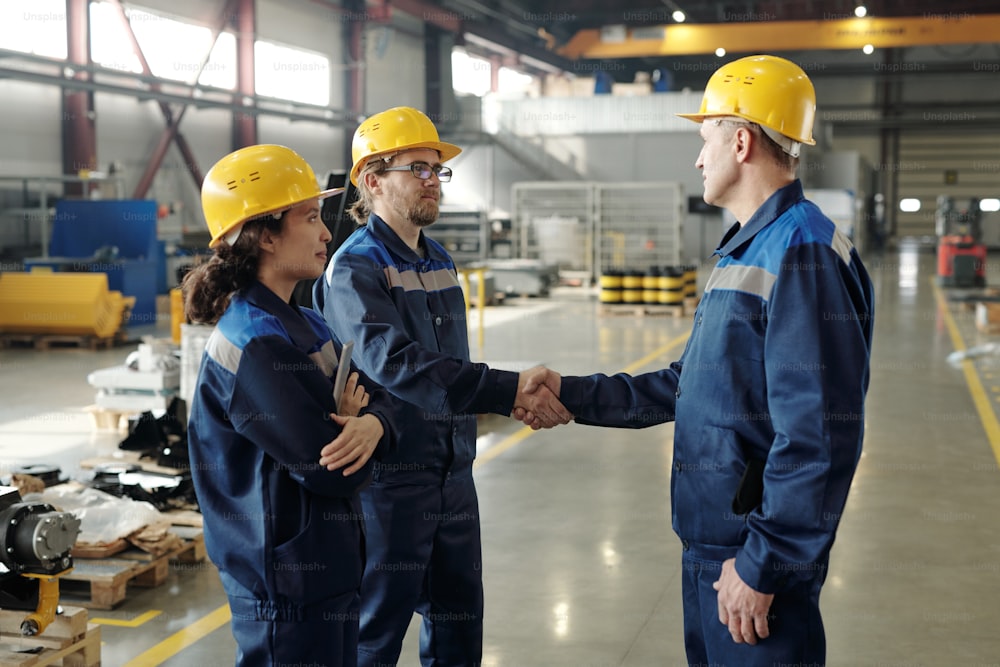Due ingegneri maschi in elmetti e abbigliamento da lavoro che si salutano con una stretta di mano in officina con una collega in piedi vicino