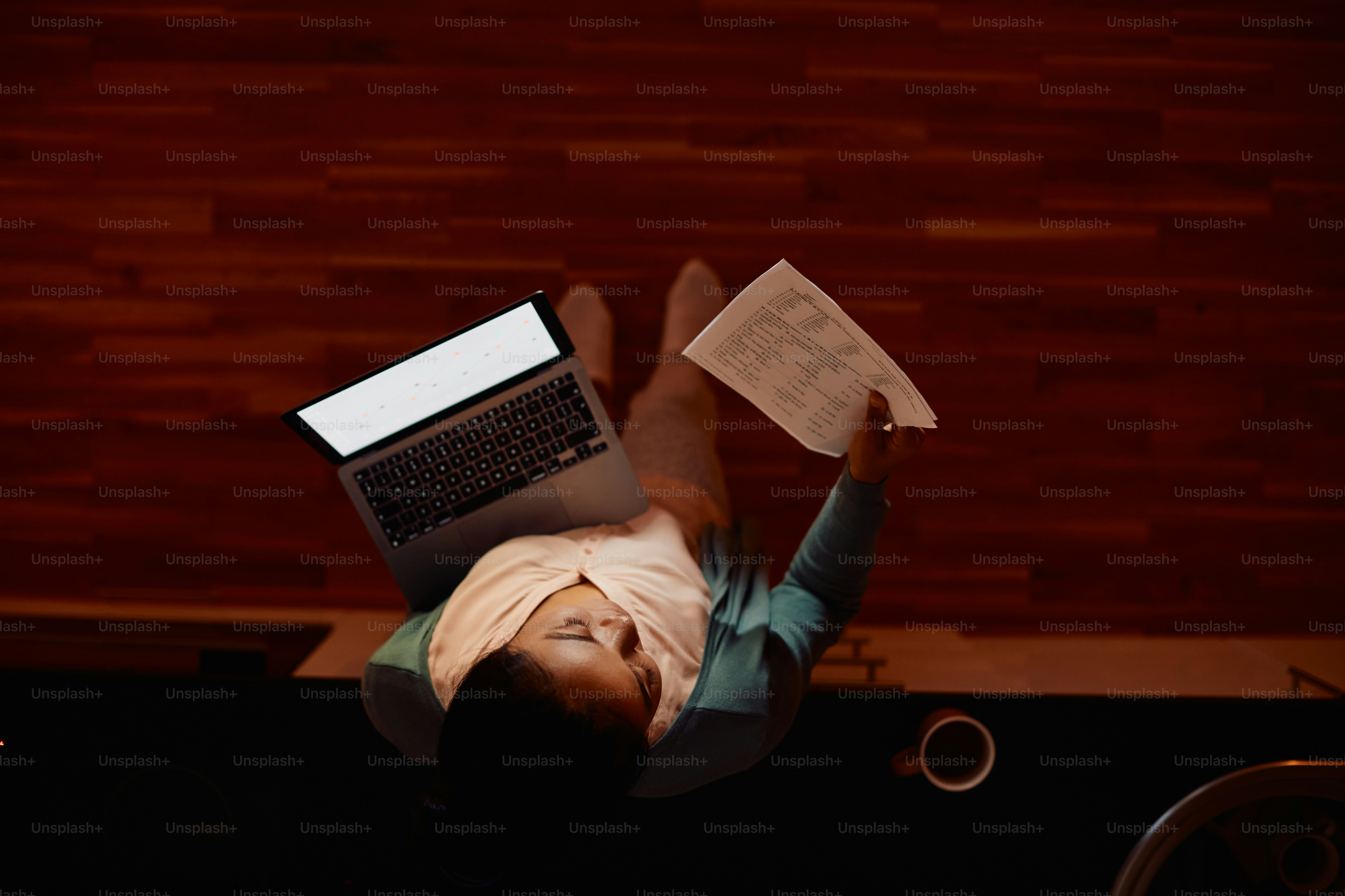 Sudut pandang tinggi pengusaha wanita menganalisis dokumen saat mengerjakan laptop di malam hari di rumah. Salin ruang.