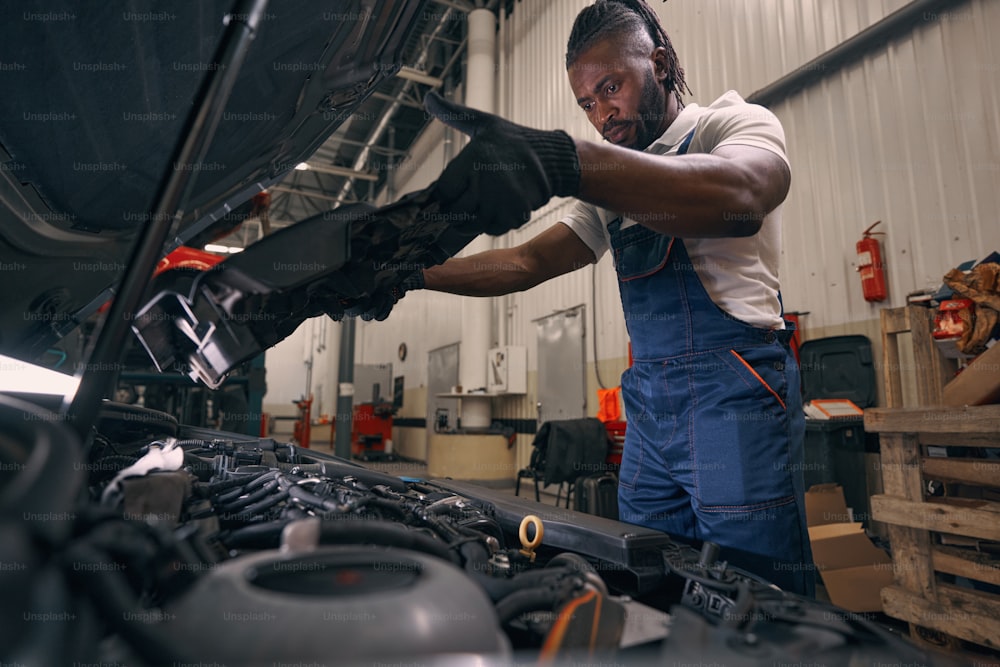 Trabajador afroamericano de un taller de automóviles sacando una cubierta de plástico negro de debajo del capó de un automóvil