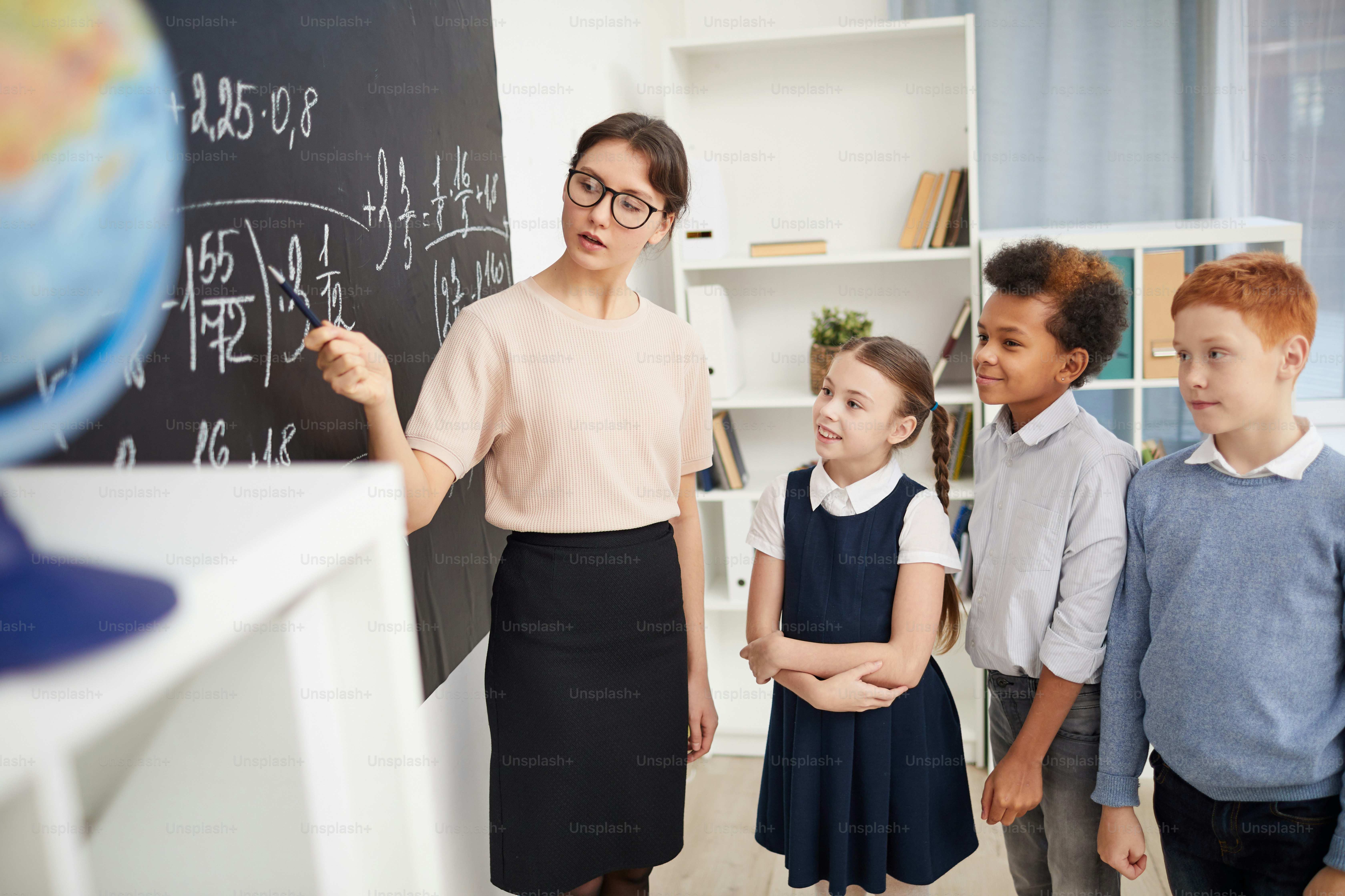 Young teacher of maths pointing at blackboard and explaining the formula to school children in the classroom