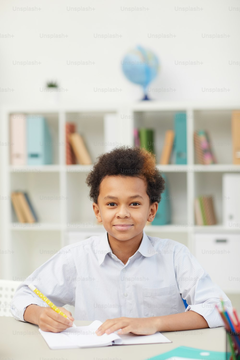 Portrait of African schoolboy sitting at desk with pen and notebook and looking at camera in the classroom