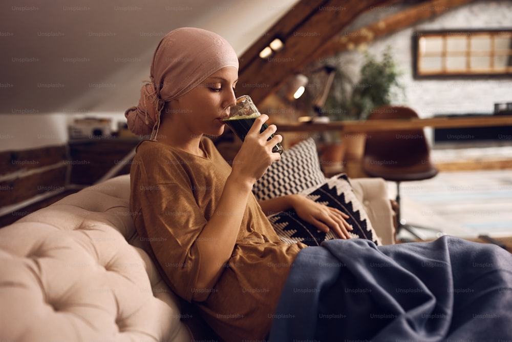 Mujer joven enferma de cáncer que toma una bebida de desintoxicación saludable mientras se relaja en el sofá de su casa.