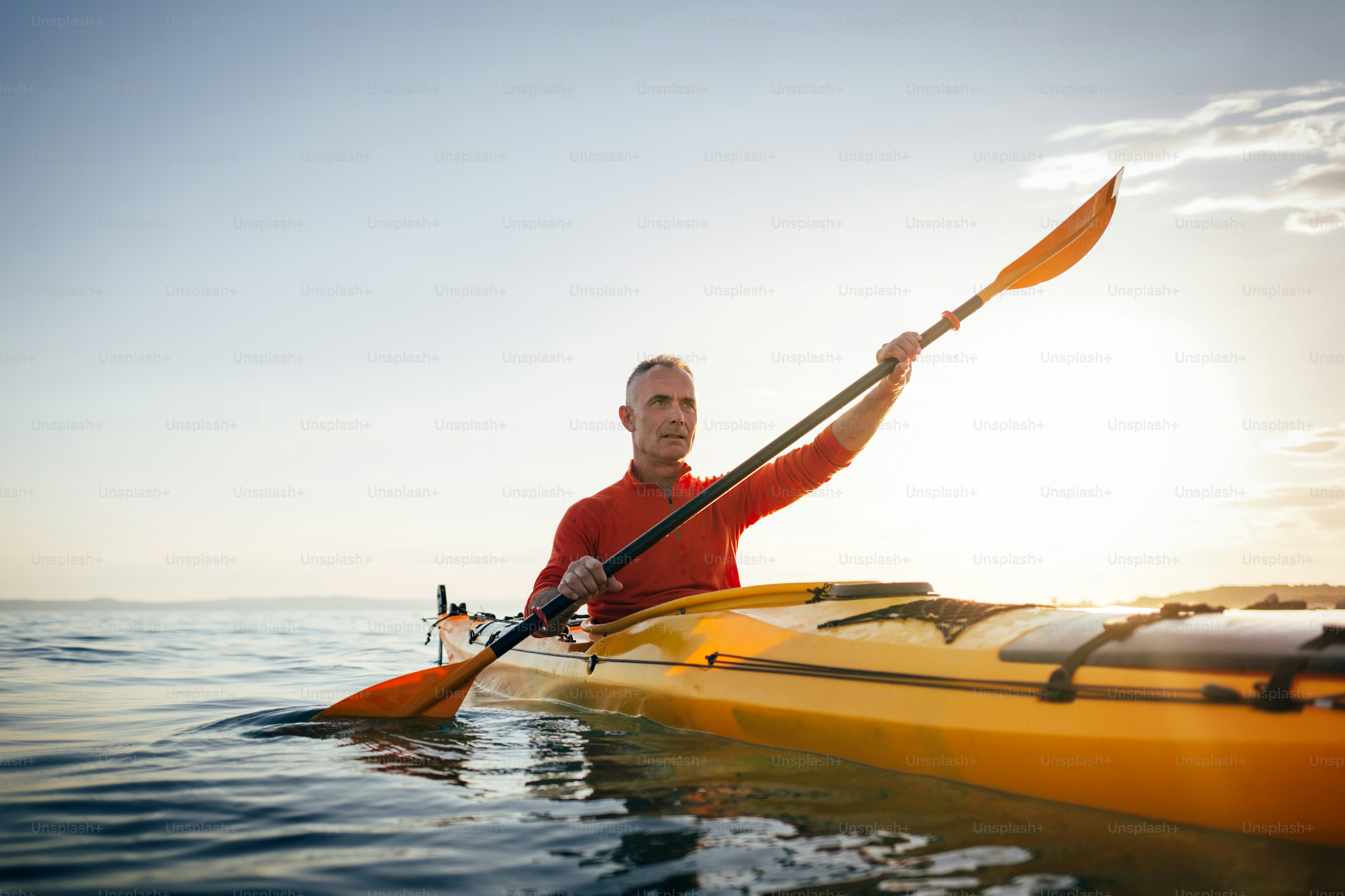 Homme âgé actif pagayant kayak de randonnée sur la mer au coucher du soleil.