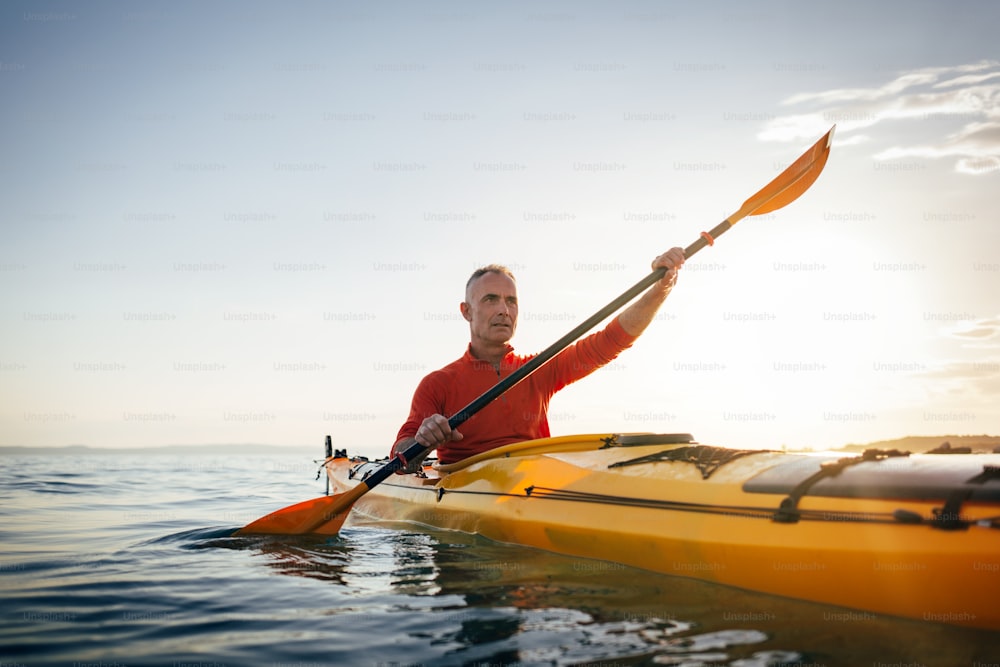 Active senior man paddling touring kayak on sunset sea.