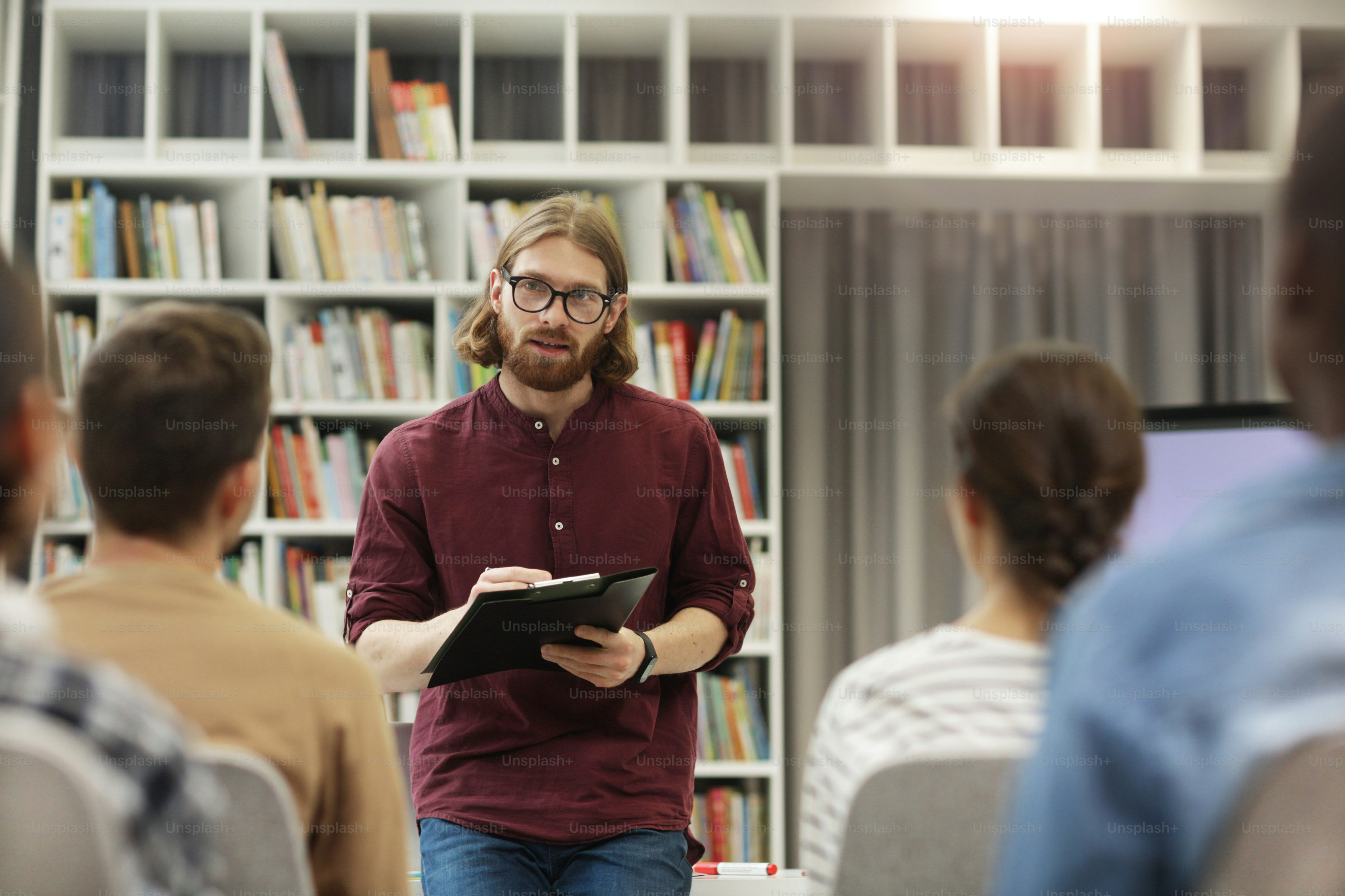 Young teacher in eyeglasses standing and talking to the students in the university library