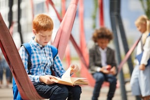 Écolier avec un sac à dos derrière le dos assis sur la balançoire et lisant un livre dans la cour de récréation de l’école