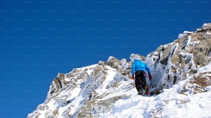 Un esquiador de travesía masculino caminando hacia una cumbre alpina remota bajo un cielo azul con cristales de hielo congelados y formaciones rocosas en el camino