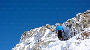 A male backcountry skier hiking towards a remote alpine summit under a blue sky with frozen ice crystals and rock formations along the way