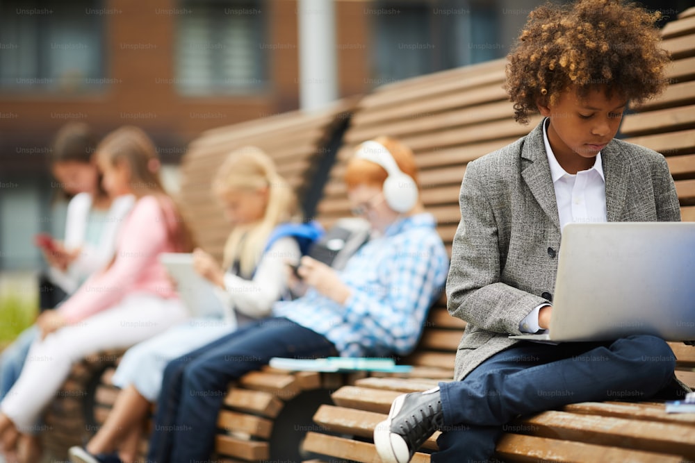 Escolar africano sentado en el banco y usando una computadora portátil para estudiar con sus compañeros de clase sentados en el fondo