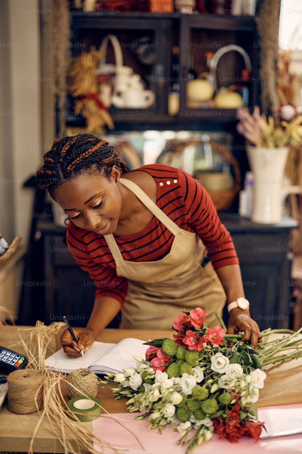 Smiling African American florist taking notes while working with fresh flowers at her workshop.