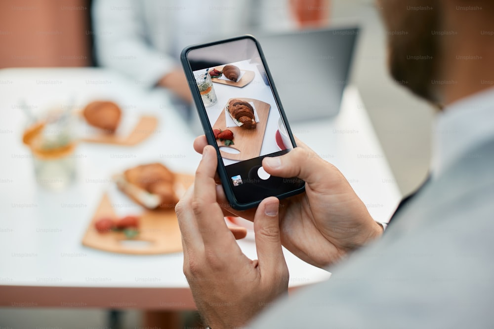 Close up of entrepreneur using cell phone and photographing his food while eating croissant for breakfast in a cafe.