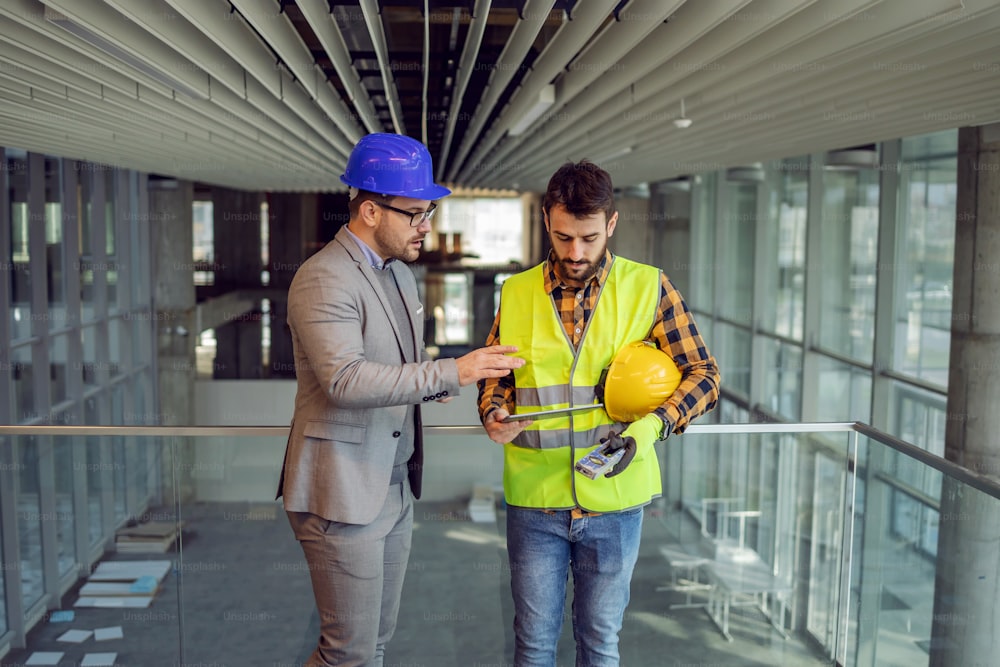 Architecte et ouvrier de la construction debout dans le bâtiment en cours de construction et regardant des plans sur une tablette.