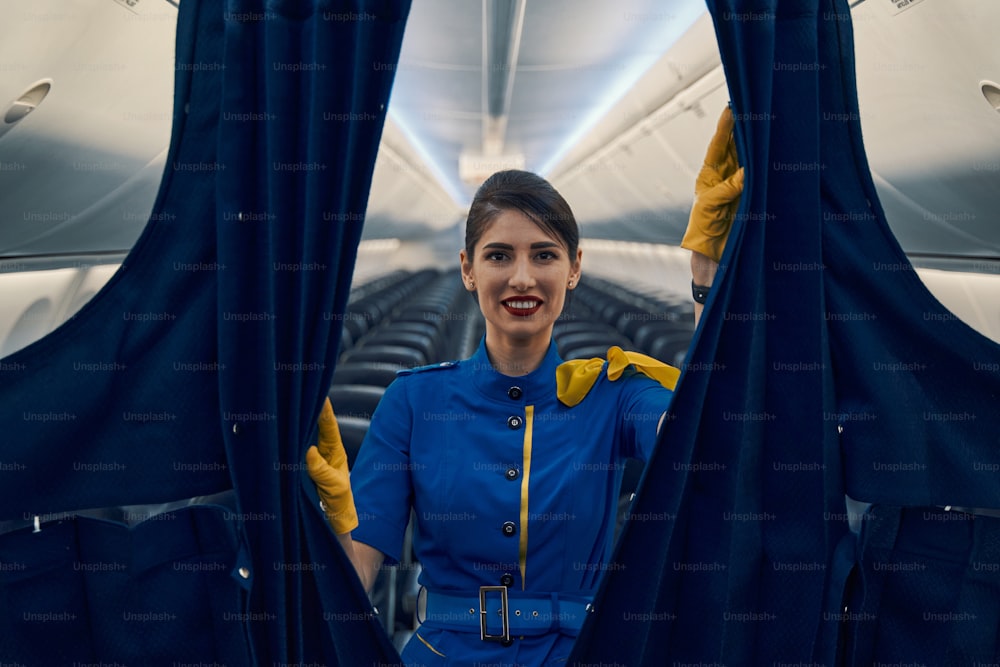 Frontansicht einer fröhlichen, attraktiven dunkelhaarigen Flugbegleiterin, die Passagierzonen in der Flugzeugkabine trennt