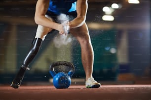 Paralympic competitor with handicapped leg standing on arena and applying powder on palms before lifting heavy kettlebell