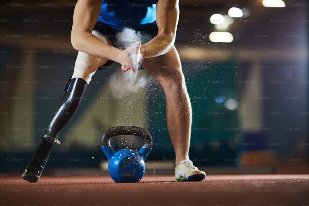 Paralympic competitor with handicapped leg standing on arena and applying powder on palms before lifting heavy kettlebell