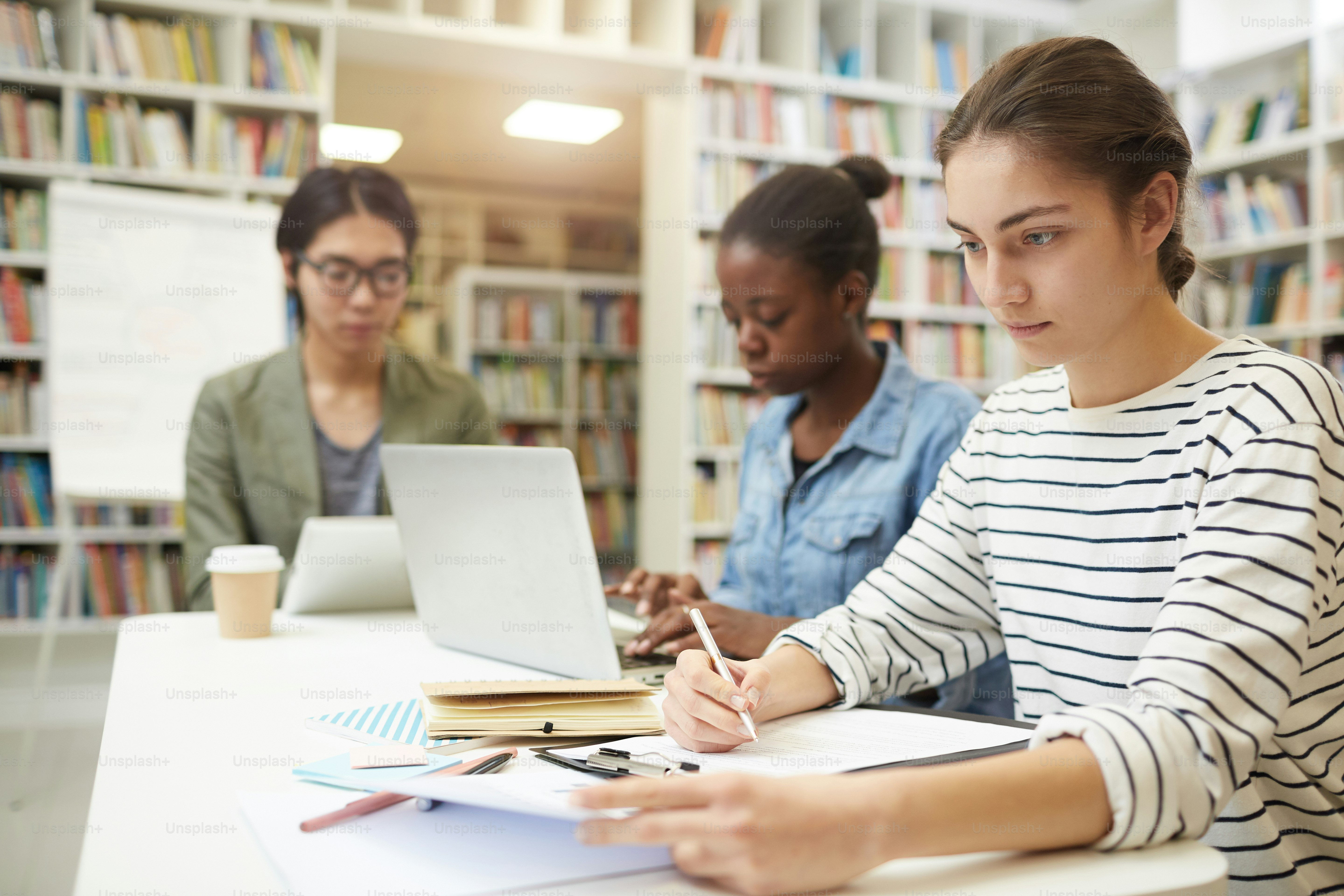 Young woman sitting at the table and working with papers she preparing for exams together with her friends in the background