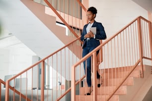 Afroamerikanische Geschäftsfrau mit digitalem Tablet, während sie bei der Arbeit die Treppe hinuntergeht. Speicherplatz kopieren.