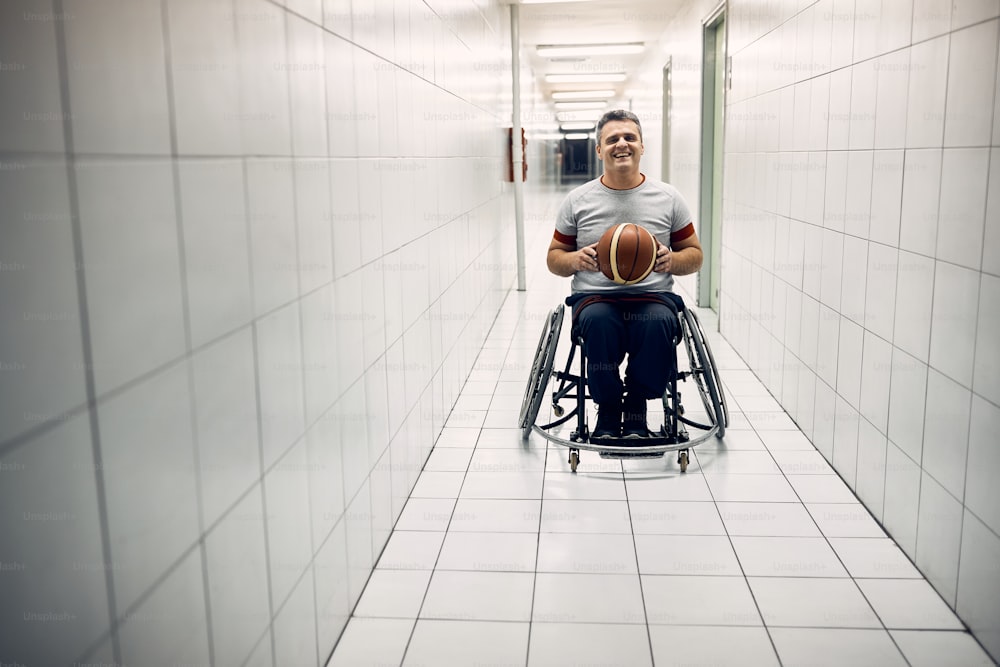 Jugador de baloncesto feliz sosteniendo una pelota en el pasillo del pabellón deportivo y mirando a la cámara.