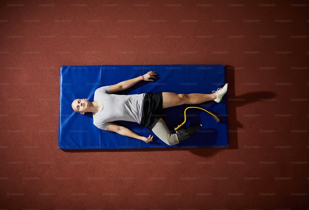 Vue d’ensemble d’un jeune compétiteur des jeux paralympiques allongé sur un tapis bleu sur le sol et se relaxant avant la compétition