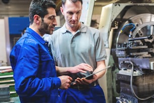 Dos trabajadores discutiendo un proyecto frente a la máquina de torno CNC escribiendo en el portapapeles