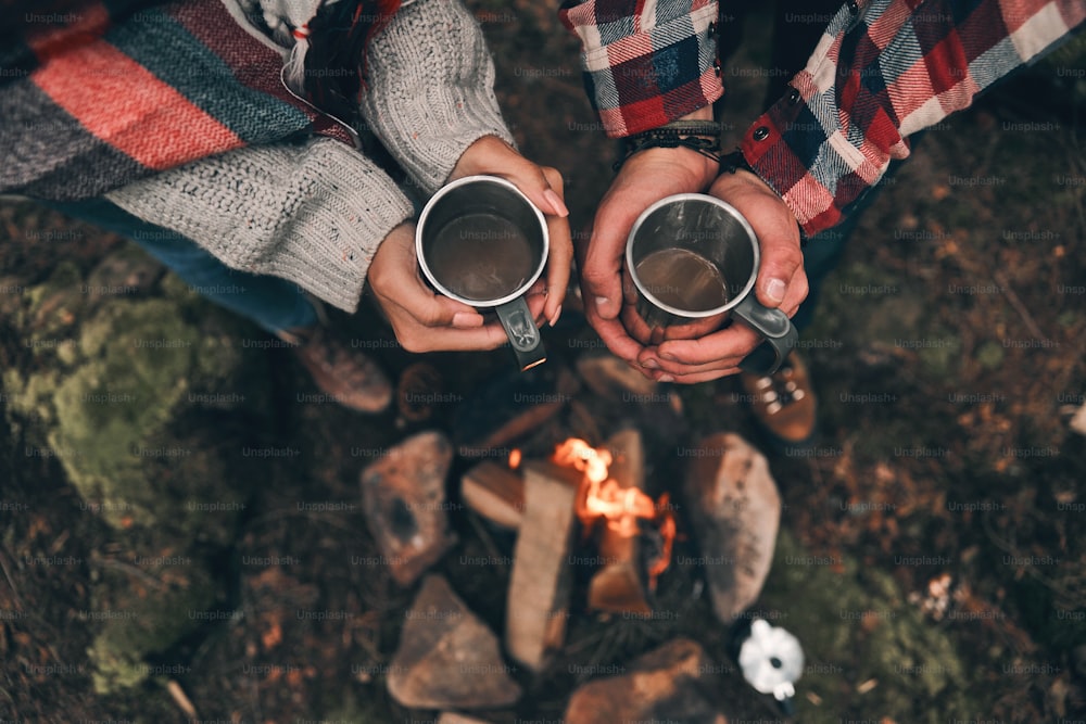 Vue rapprochée de dessus d’un jeune couple tenant des tasses tout en se réchauffant près du feu de camp
