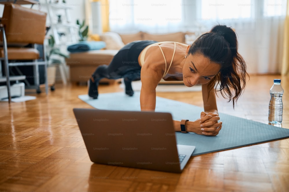 Desportista adulta média usando laptop enquanto se exercita em pose de prancha em casa.