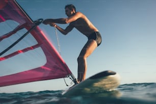 Vista ad angolo basso della silhouette del surfista in equilibrio sulla tavola da windsurf. Windsurfer uplift vela per la navigazione in windsurf sul mare al tramonto.