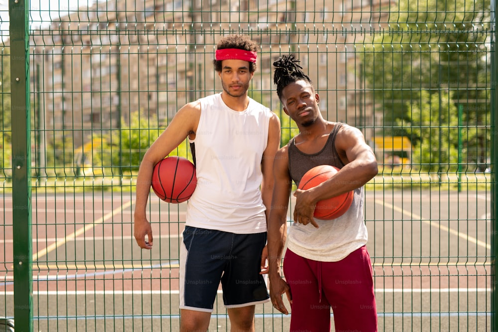 Small group of two young professional basketballers in activewear standing by fence surrounding court