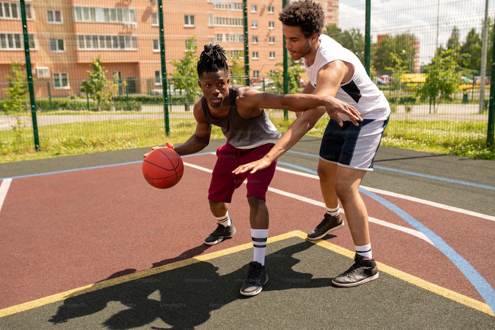 Jeune instructeur de basket-ball aidant un sportif africain avec l’un des exercices lors d’un entraînement en plein air sur le terrain