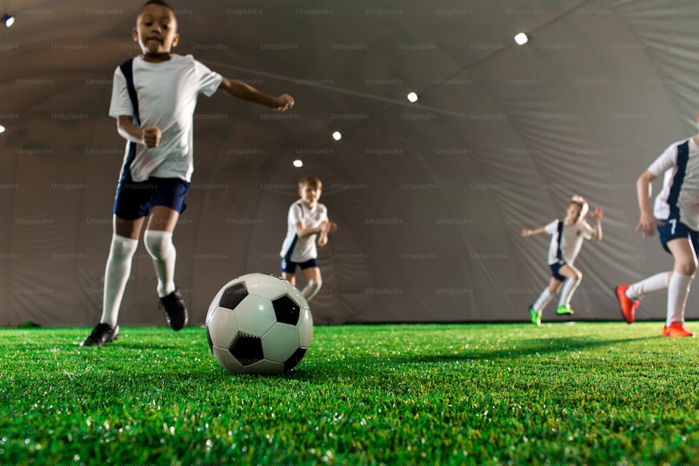 Bola de futebol no campo verde e pequenos jogadores correndo em direção a ela durante o jogo