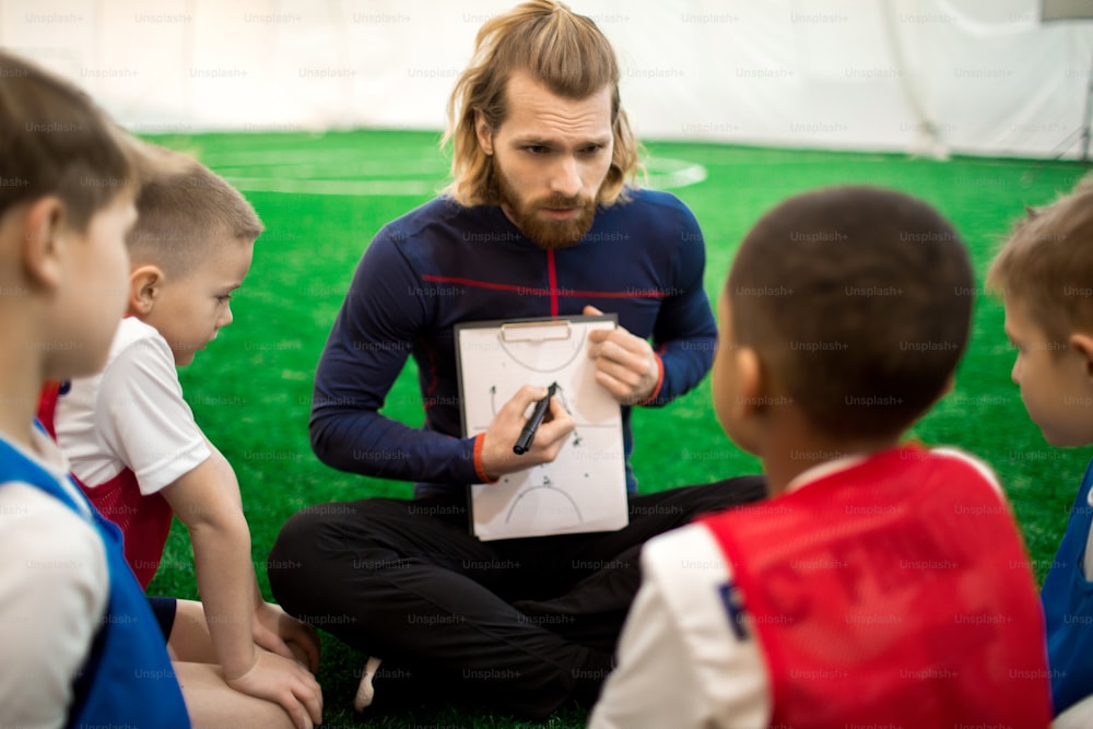 Confident football trainer drawing scheme of moving on pitch during game and explaining it to youhful players