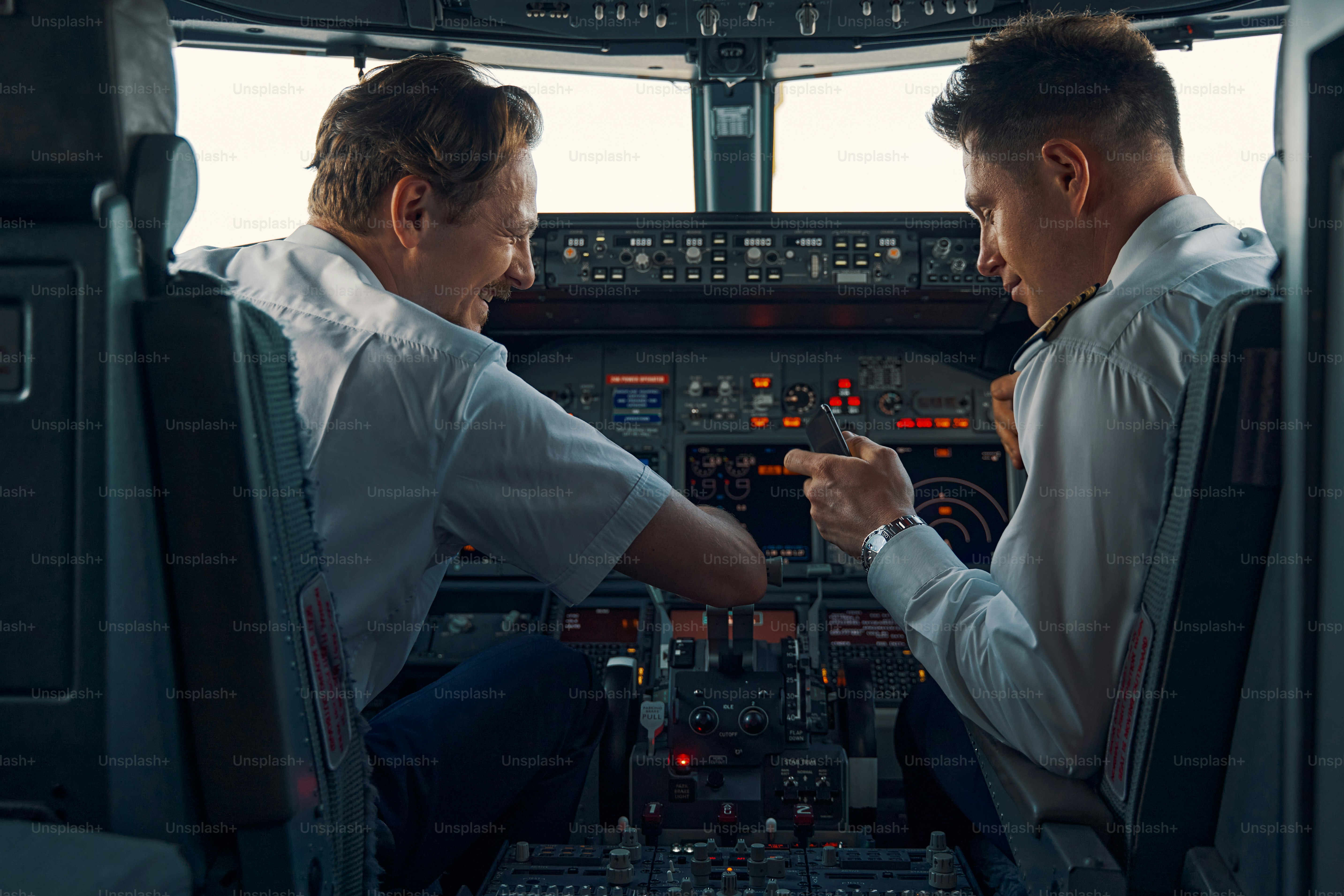Back view of an airline captain and a first officer with a smartphone in his hand sitting in the cockpit