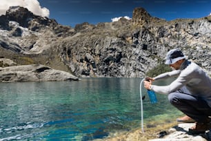 male backpacker dressed in gray filters drinking water from a turquoise mountain lake in the Cordillera Blanca in the Andes in Peru